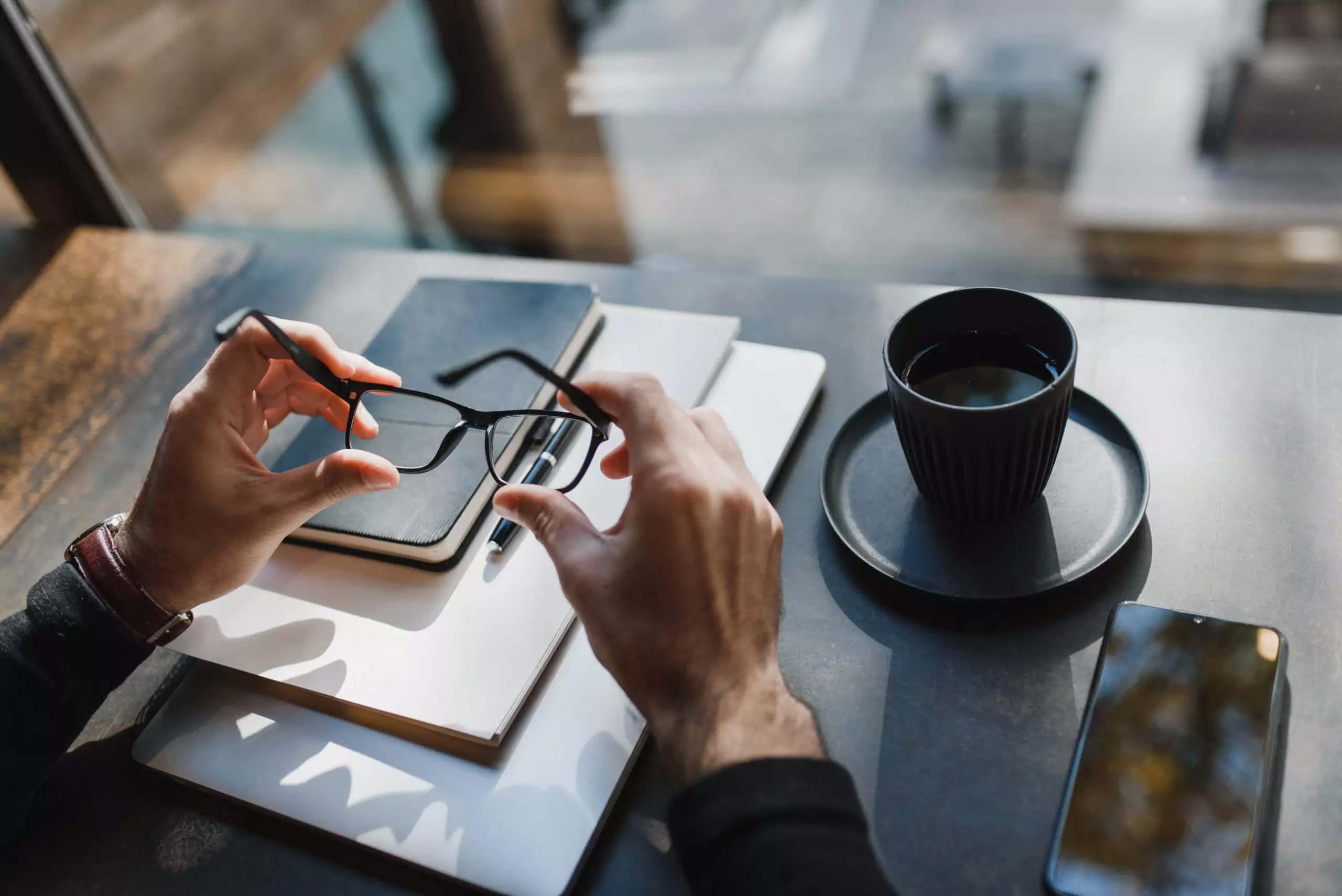 A man holding glasses above his notebooks and next to a cup of coffee and his phone