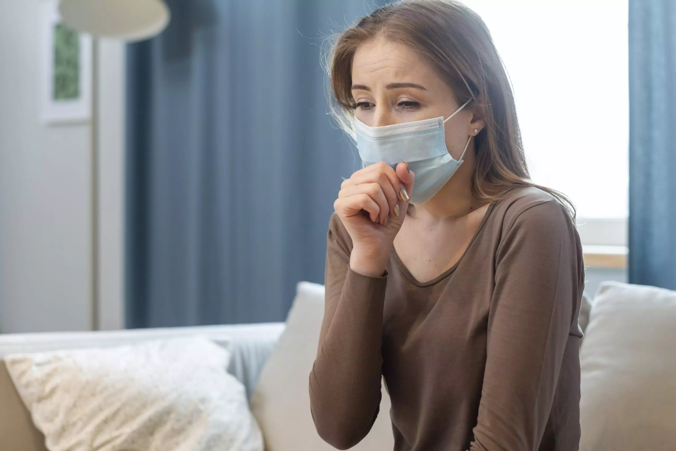 Woman sitting on sofa with mask on and coughing from long COVID